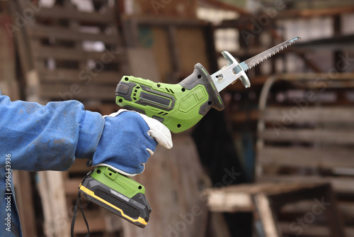 A craftsman in a protective glove holds a compact green cordless reciprocating saw. The tool is designed for fast and efficient sawing of wood, metal, plastic and plywood. Power tool.