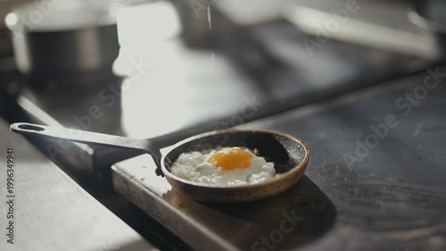 Soft light glows over a tiny pan. Fried egg rests gently in the skillet. Wooden board holds the pan. Warm, rustic kitchen setting. Ideal for food or lifestyle content. No people or animals visible