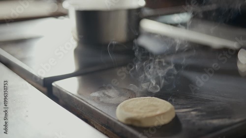 Hands place round bread on hot griddle. Steam rises from sizzling surface. Cooking scene suggests morning meal prep. Crisp texture promised by heat. Natural kitchen lighting adds warmth