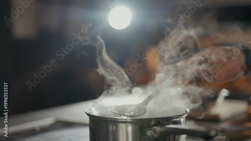 Steam rises from a steaming pot on a stove. Hand gently stirs the contents. The chef's tattooed arm adds a bold touch. Warmth and aroma fill the kitchen air. Perfect for culinary or cooking scenes
