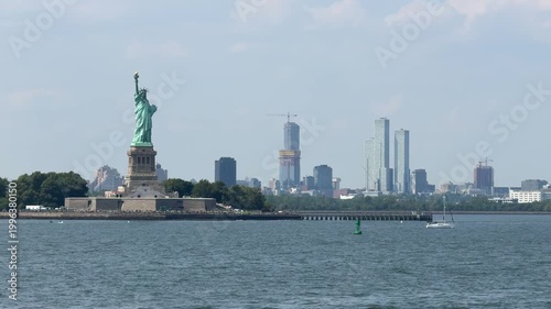 The Statue of Liberty stands on Liberty Island with New York Harbor waters and distant city skyline behind it. Iconic American landmark represents freedom, travel, and tourism under clear blue sky.