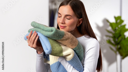 Foto Young Caucasian white woman holding and smelling clean sweaters on light gray wardrobe in home background