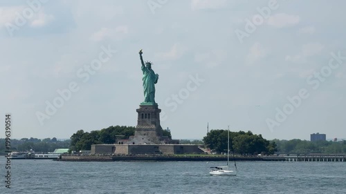 The Statue of Liberty stands tall on Liberty Island while a white sailboat moves across the harbor water. Bright summer daylight creates a scenic travel and tourism view of New York City.