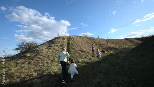 A Happy Family with Children Enjoys a Scenic Walk Uphill in Nature under a Bright Blue Sky on a Sunny Day, Exploring the Outdoors Together