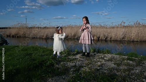 Two Joyful Little Girls Play with a Dandelion by a Serene River, Blowing Its Seeds into the Clear Blue Sky on a Beautiful Sunny Day, Capturing Moments of Carefree Childhood in Nature