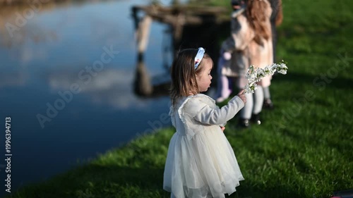 Adorable Little Girl in White Dress Holds Delicate White Flowers by the Tranquil Riverside, Enjoying a Sunny Spring Day in Nature with Family