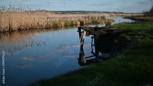 A Serene River Landscape with a Rustic Wooden Pier and Golden Reeds Reflecting the Blue Sky and Clouds on a Calm Day