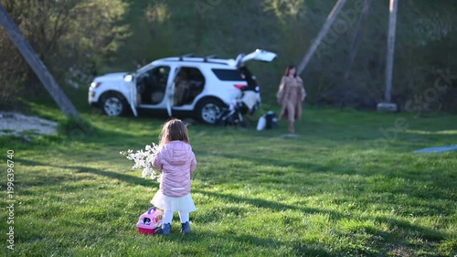 Little Girl Holding White Spring Flowers and a Pink Bag in a Green Field, with a Family Car and Adult in the Background, Enjoying a Peaceful Outdoor Moment
