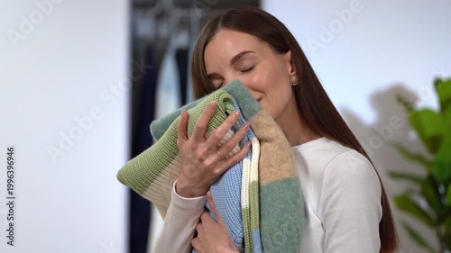 Young smiling woman pressing soft wool sweater to cheek, smelling aroma. Wardrobe, green plant on home interior background. Concept of fresh laundry routine, tactile comfort, knitwear clothing care