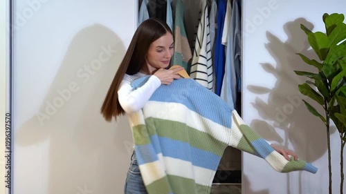 Young woman hugs blue striped sweater on hanger, smiling and twirling near open wardrobe at home. Concept of laundry freshness, soft knit comfort, aromatic fabric care and cozy domestic lifestyle