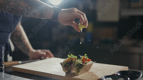 Hands season tacos with care. Tattooed arms move with precision. Food rests on a wooden board. Light shines from a single source. Taco preparation in a dim kitchen. Great for food, cooking