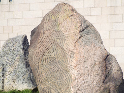 Two large boulders are intricately carved with swirling, interconnected patterns, displayed in front of a light-colored stone building wall.
