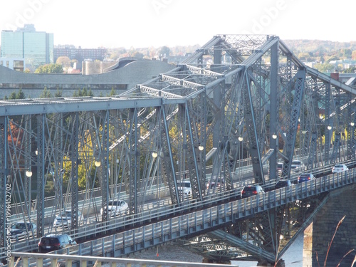 A large grey steel truss bridge spans over water with cars driving across, framed by a city skyline background.
