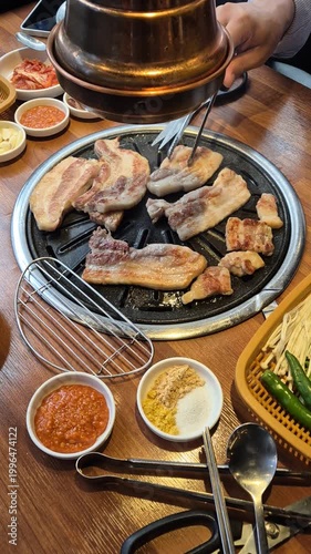 Korean barbecue pork belly grilling on tabletop grill with side dishes.