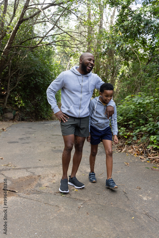 Fototapeta premium African American father and son walking on paved woodland trail laughing, wearing gray hoodies