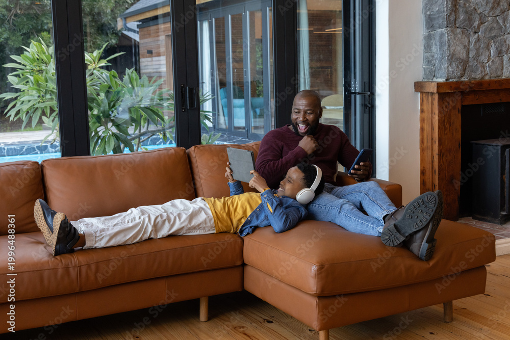 Naklejka premium African American father and son lounging at home, son using tablet while father holding phone