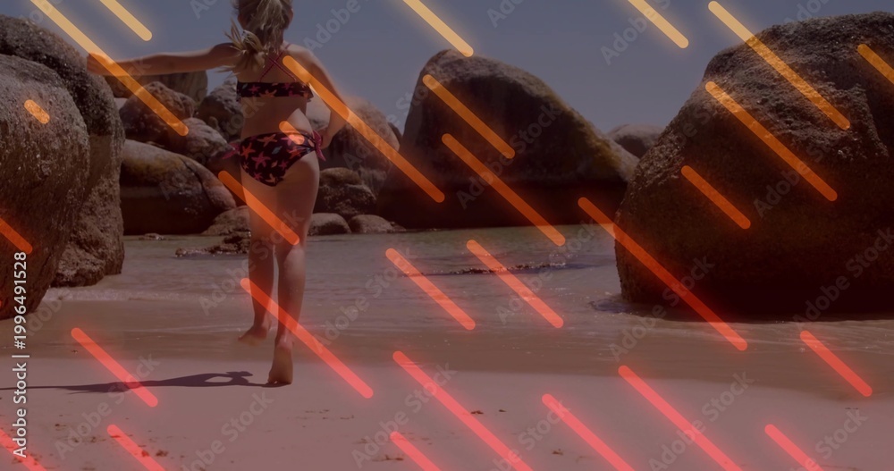 Obraz premium Wading girl in star bikini moving into shallow sea on beach large boulders neon-orange or pink strea
