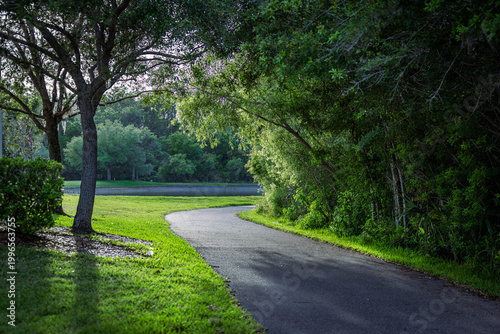 Φωτογραφία Paved walking path curving through a lush green park with trees and a pond in the distance, USA