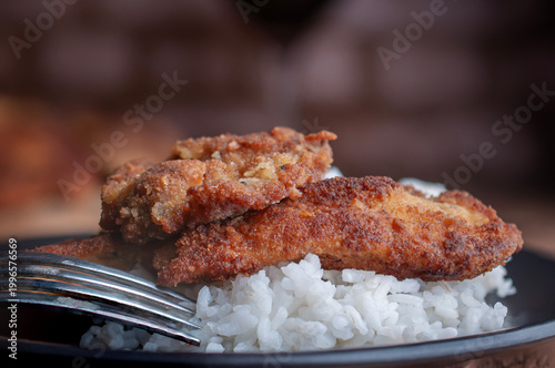 Milanesas de pollo pescado o carne de res sobre arroz blanco al vapor con un tenedor al lado, sobre un plato negro y pared de ladrillos al fondo, macro o acercamiento.