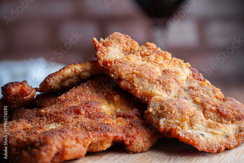 Milanesas de pollo pescado o de res sobre una tabla de cortar con un tazon de arroz blanco al vapor, una copa de vino tinto fuera de foco y una pared de lacrillo al fondo.