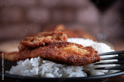 Milanesas de pollo pescado o camarones empanizados sobre arroz blanco a vapor en un plato negro con tenedor y pared de ladrillo al fondo