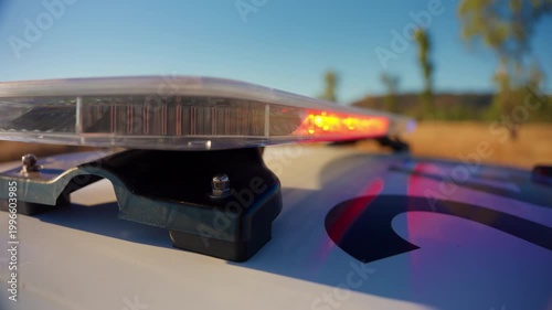 Close-up view of police car light bar with flashing red and blue emergency lights on white patrol vehicle roof against clear blue sky in rural outdoor setting with trees and golden grass.
