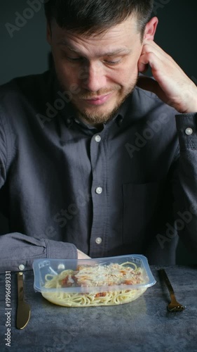 Melancholic man sitting at dark table receives prepackaged pasta meal in plastic container, expressing sadness, disappointment about instant lunch delivery service. Vertical shot.