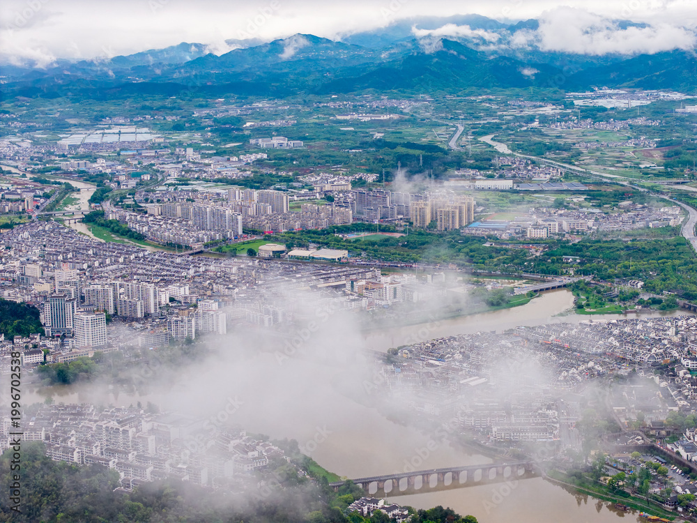 Obraz premium An aerial view captures the historic Huizhou Ancient City in She County of Anhui Province in China with mist flowing over the river and traditional buildings surrounded by mountains