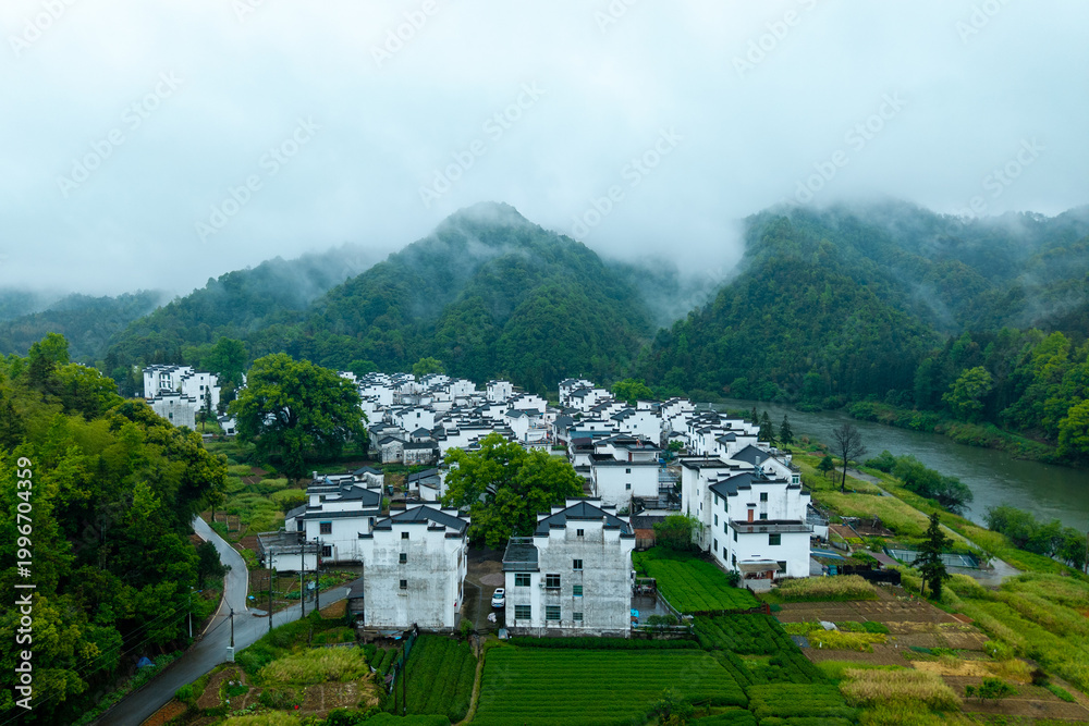 Fototapeta premium Aerial landscape of Huizhou Ancient City, She County, Anhui, China, displaying ancient white houses and green fields by a river amidst misty mountains