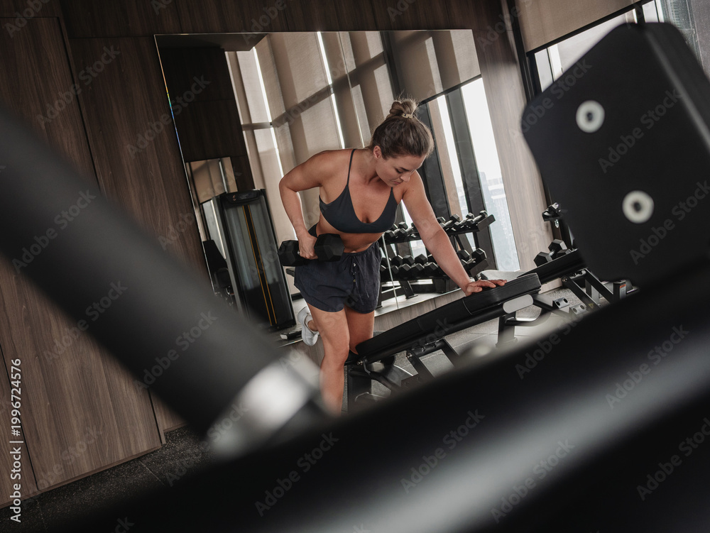 Fototapeta premium Athletic woman performing one arm dumbbell row on bench in modern gym