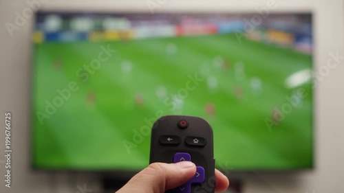 Person changing channels with a roku remote control while watching a live soccer game on a smart television, relaxing in a living room and enjoying free time at home