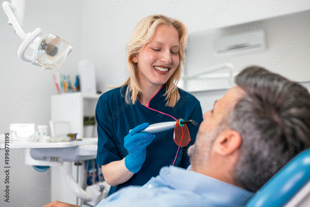 Fototapeta premium Female dentist using dental curing light on male patient. Professional teeth whitening, filling procedure, and healthcare concept. Woman doctor performing treatment in modern stomatology clinic room.