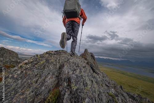Fotografía Male hiker with backpack and trekking poles climbing a sharp rocky peak in the mountains under a dramatic sky