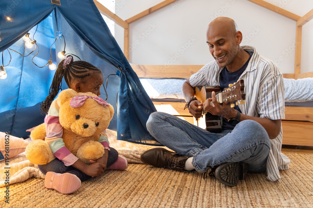 Fototapeta premium Indian dad and African American daughter sitting on bedroom rug, playing guitar, she hugging teddy