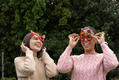 Diverse female friends wearing knit sweaters laughing while adjusting holiday glasses at hedge