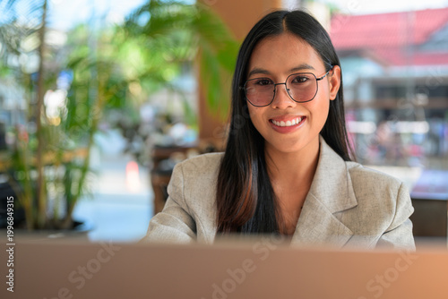 A smiling Asian woman from Thai Isan looking at camera wearing glasses while working on a laptop in a professional cafe.