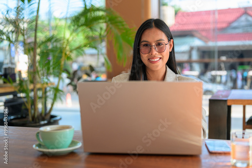 A smiling Asian woman from Thai Isan looking at camera wearing glasses while working on a laptop in a professional cafe.