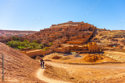 Visitors walk along a path towards Ait Benhaddou in Morocco. The village showcases traditional earthen clay architecture against a clear blue sky. This site is a UNESCO World Heritage Site.