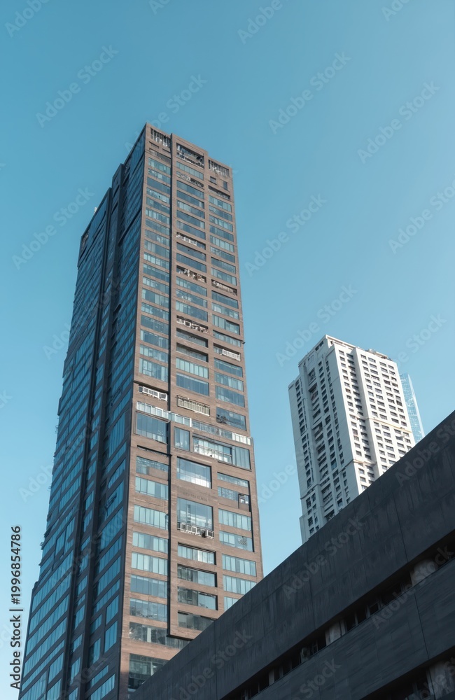 Obraz premium Tall dark skyscraper next to white residential tower under clear blue sky. Modern glass buildings rise high above city streets. Urban architecture cityscape view from below.