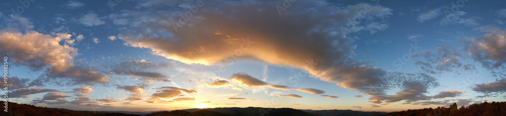 Obraz premium Stunning sunset fills sky with rich hues of blue and orange. Dramatic clouds with hints of pink and gold spread across sky. Silhouettes of hills in foreground add depth to breathtaking evening scene.