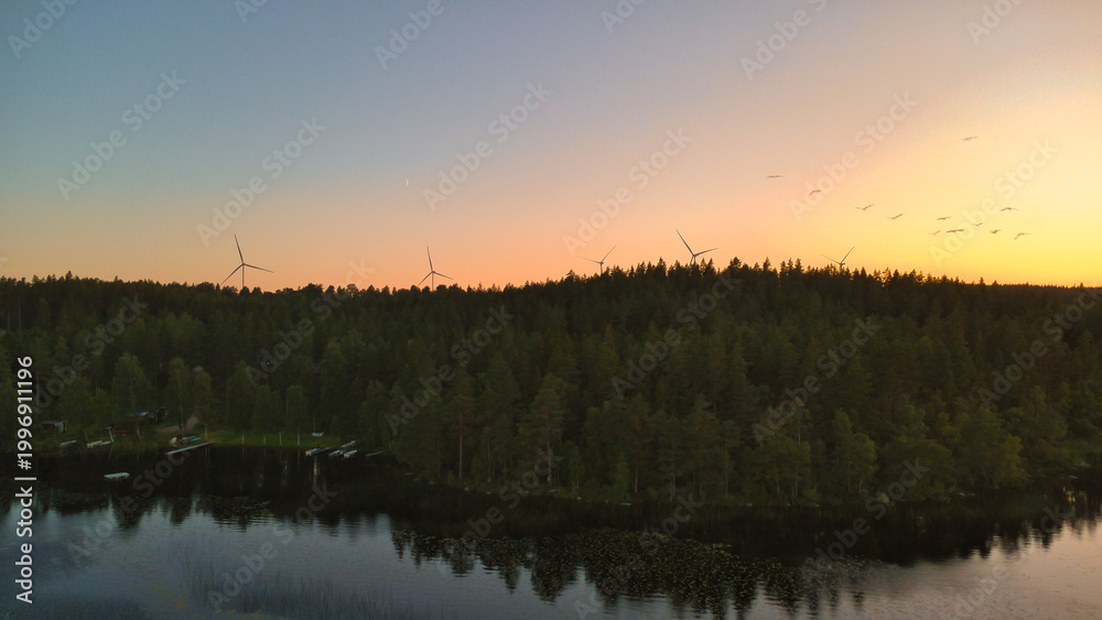Obraz premium Wind Turbines Above a Forest Near a Lake at Sunset with Birds in the Sky