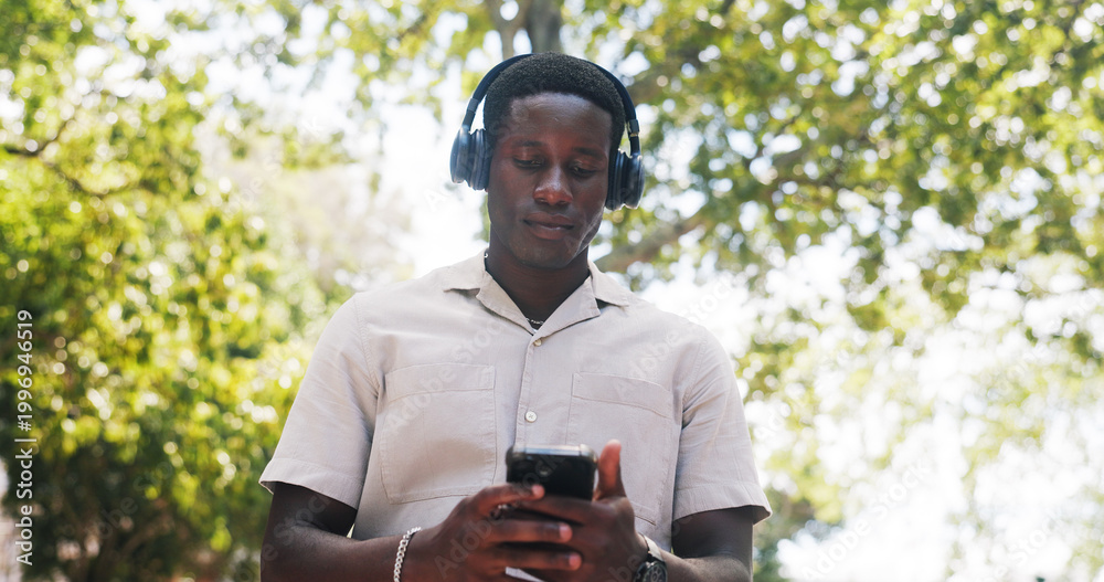 Fototapeta premium Black man, phone and music at park with headphones, streaming or listen to sound with low angle in summer. African person, scroll and outdoor with mobile app, texting or audio subscription in nature