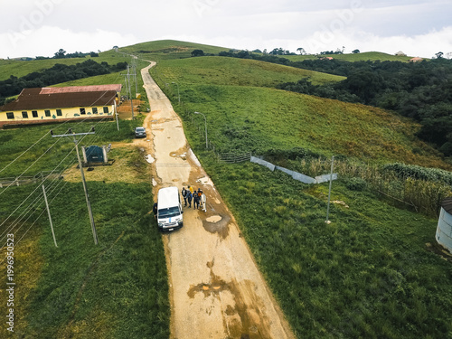 Aerial view of a winding dirt road through lush green hills, past a yellow-brown building and a white van with people, Obudu, Cross River, Nigeria.
