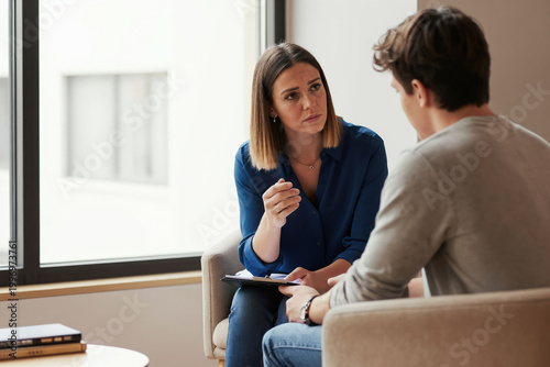 Female therapist talks to teenage boy in office about his problems. Woman listens attentively, offers support and guidance. Teenager looks troubled, seeking help. Professional mental health session.