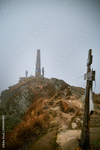Hikers reach the summit with a tall stone monument on a foggy day in the mountains. Autumn hiking in Carpathian Mountains, Ukraine