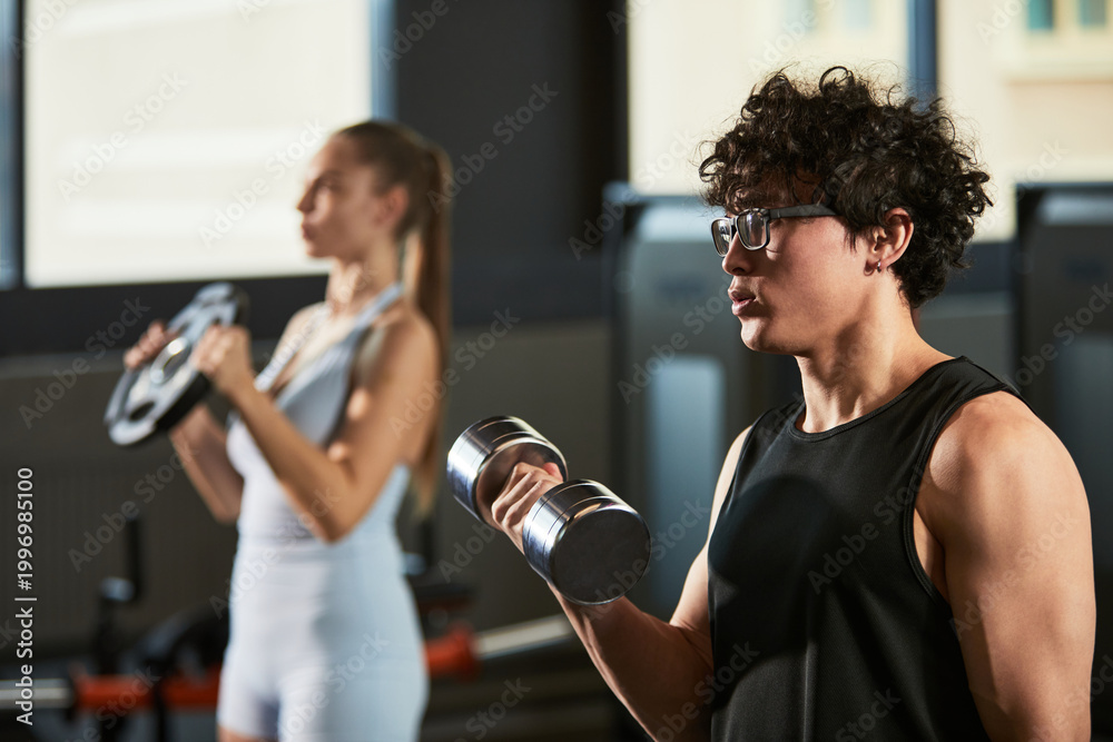 Fototapeta premium Young adult man lifting dumbbell during strength workout while young adult woman training with weight plate in gym. Ideal for fitness marketing, personal training, healthy lifestyle campaigns