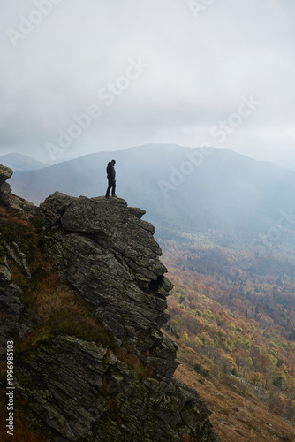 Hiker stands on rocky outcrop overlooking misty mountain landscape in autumn. Autumn hiking in Carpathian Mountains, Ukraine