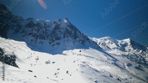 A cable car gondola glides across dramatic snow-covered alpine slopes beneath the towering Mont Blanc massif under a vivid blue sky in Chamonix, French Alps.