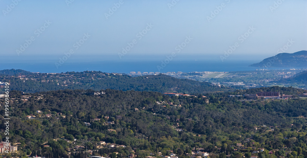 Fototapeta premium Aerial view of Grasse, pine covered hills and wooded slopes descend to the Mediterranean and the Cote d'Azur, curved bay and headlands sit under clear midday light.