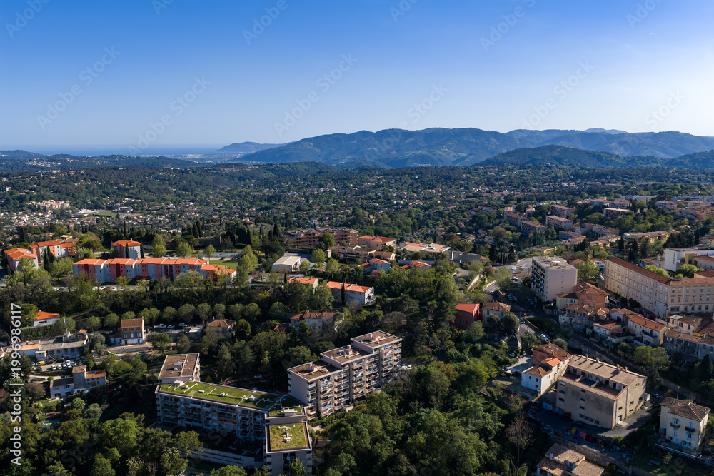 Fototapeta premium Aerial view of Grasse, France shows terracotta roofs, green rooftops, and tree lined streets across a valley, with the Prealps in midday light and rolling hills.